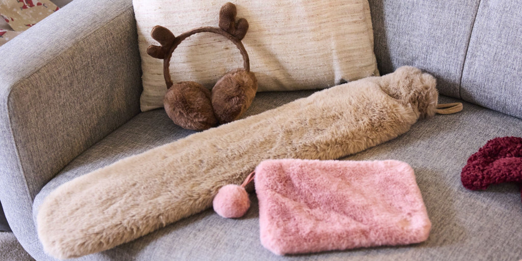 Cosy winter warmers on a grey sofa: reindeer earmuffs, a beige furry hot water bottle cover, and a pink fluffy pouch with pom-pom zip—perfect for autumn evenings and winter walks.