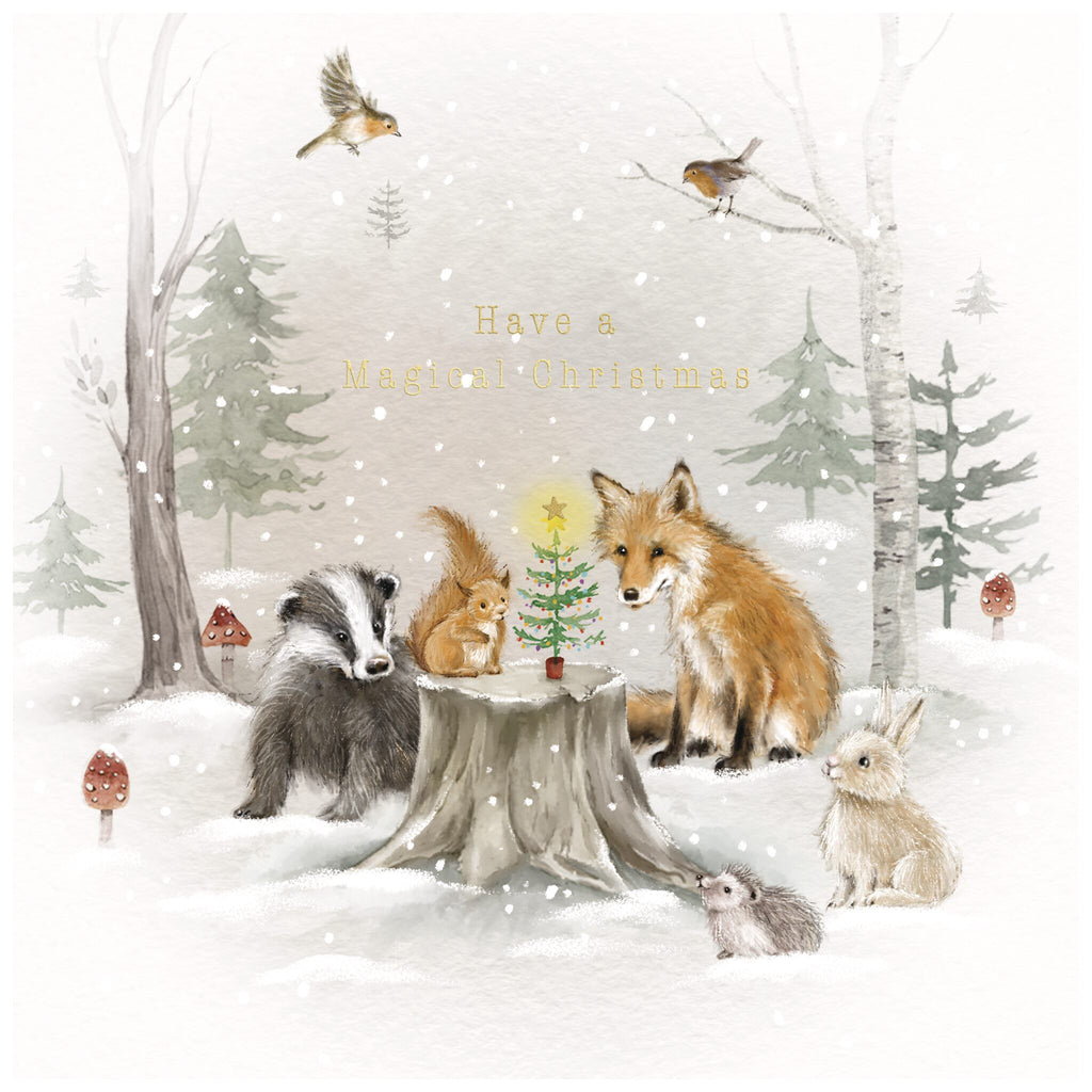 A Christmas card of a snowy woodland scene with a badger, squirrel, fox, rabbit, and mouse gathered around a tree stump topped with a small decorated Christmas tree. Two birds perch on branches above, while snow falls gently in the background. Trees and mushrooms are visible, and the text reads: 'Have a Magical Christmas'.