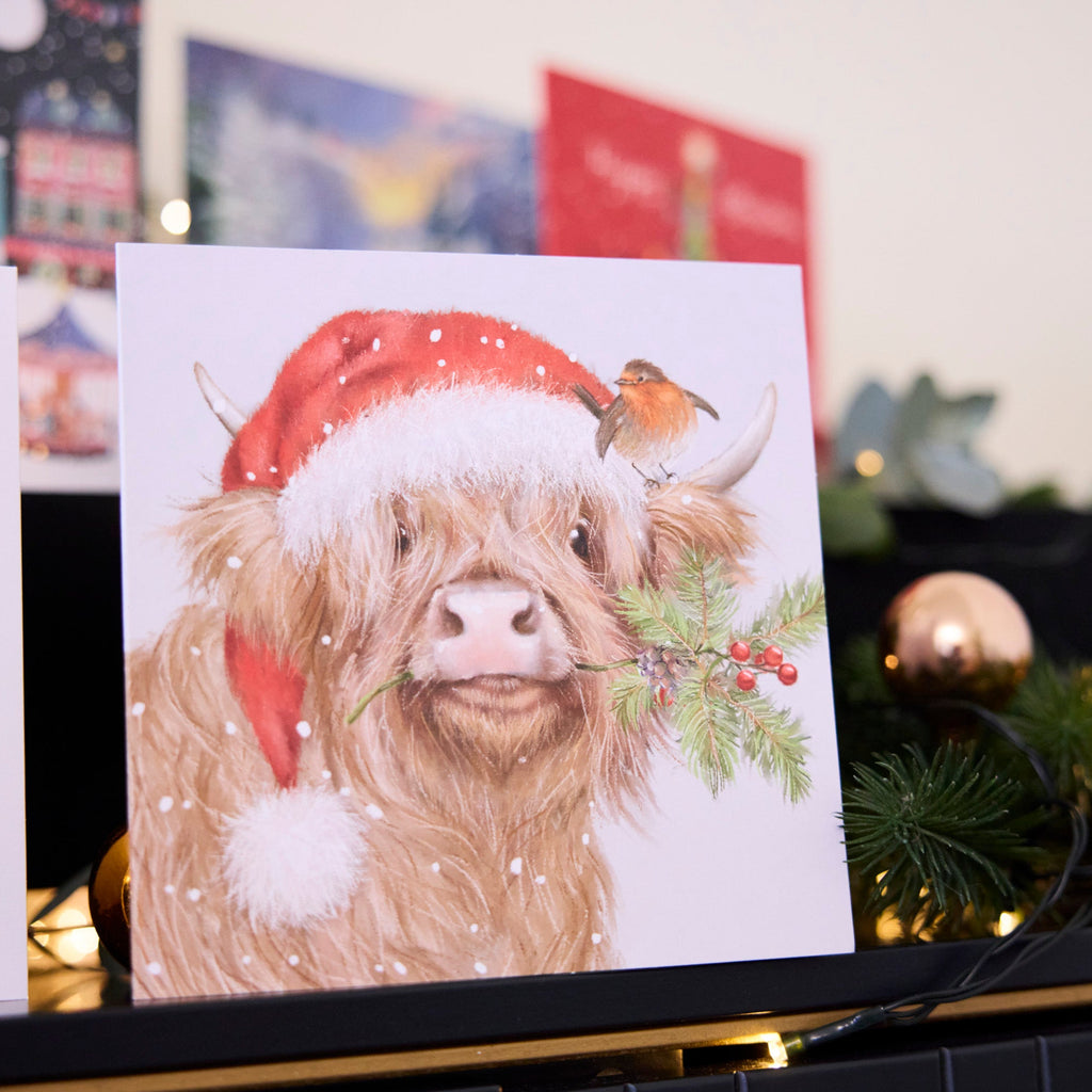 Clover the Highland Cow Christmas card is sitting on top of a mantlepiece. 