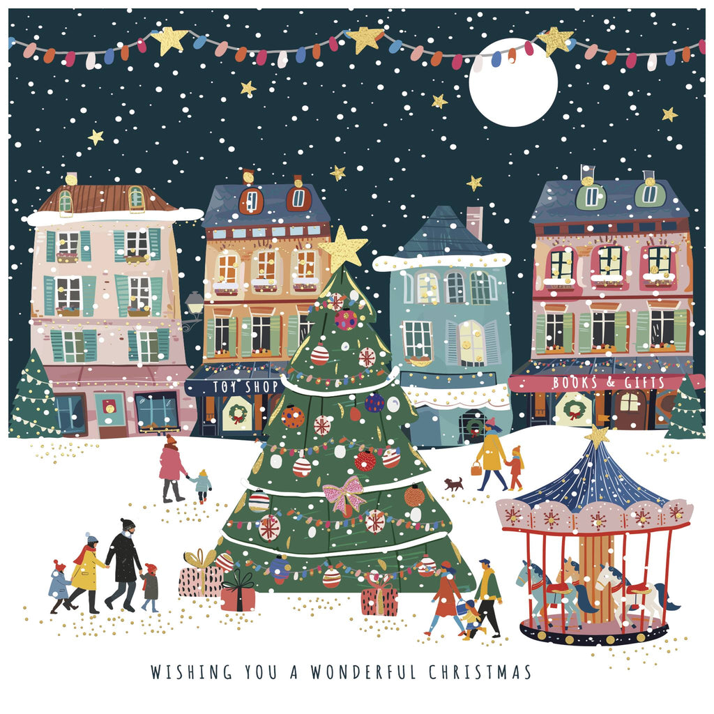A Christmas card of a festive town square scene with a large decorated Christmas tree in the centre, surrounded by people walking, children playing, and some carrying gifts. Colourful buildings in the background include shops labelled 'TOY SHOP' and 'BOOKS & GIFTS'. A carousel with children riding it appears on the right, while snowflakes, stars, and a full moon fill the dark sky. The text at the bottom reads: 'WISHING YOU A WONDERFUL CHRISTMAS'.