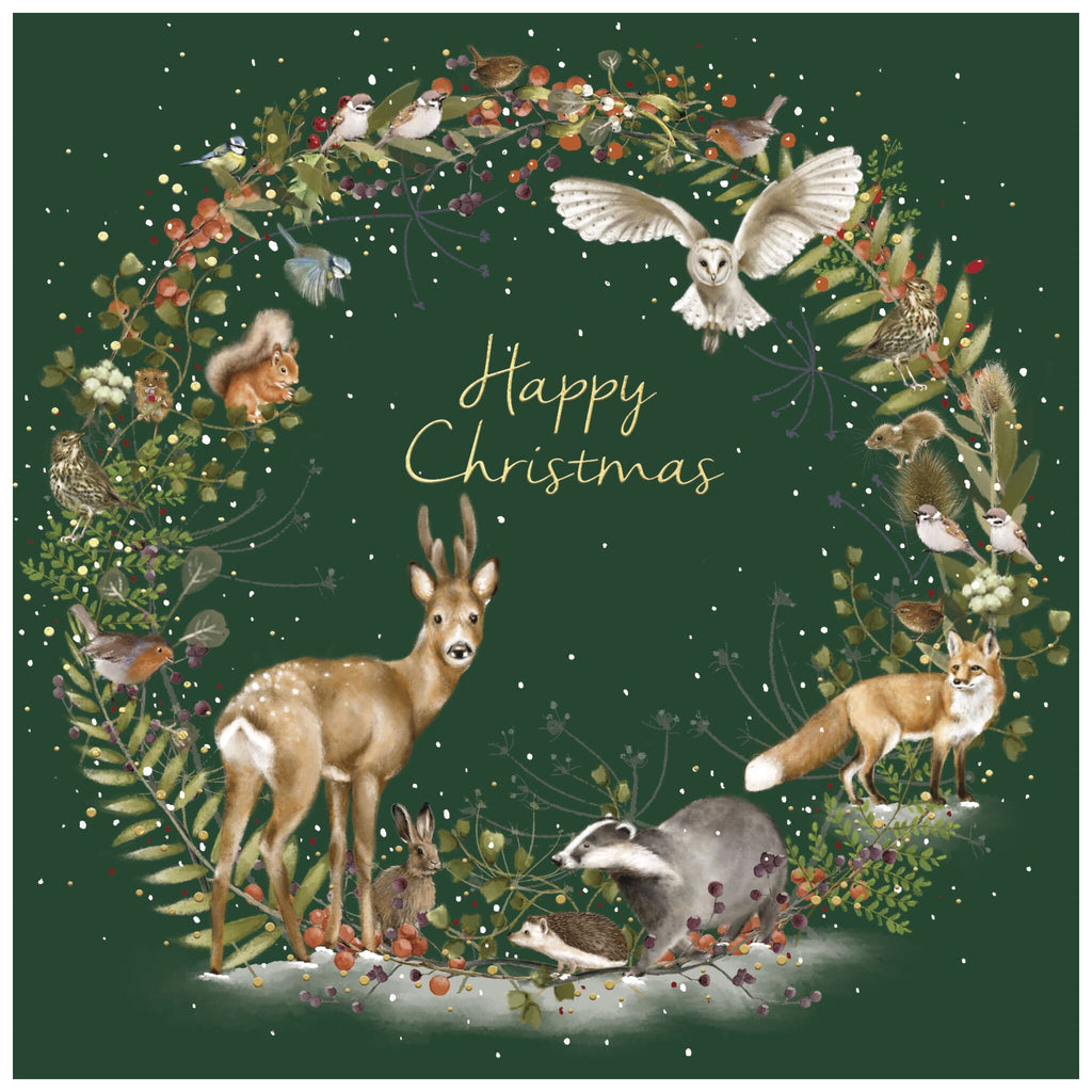 A Christmas card of a woodland scene featuring a circular wreath made of leaves, berries, and flowers on a dark green background, with snowflakes scattered throughout. Inside the wreath are a deer, fox, badger, hedgehog, rabbit, squirrel, owl in flight, and various birds perched on branches. In the centre, the text 'Happy Christmas' is written in gold cursive lettering.