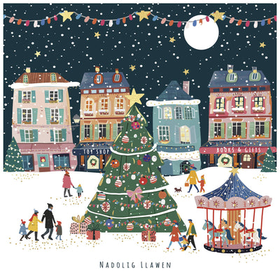Illustration of a festive town square with a Christmas tree, carousel, and people walking around. The Welsh greeting "Nadolig Llawen" appears at the bottom.