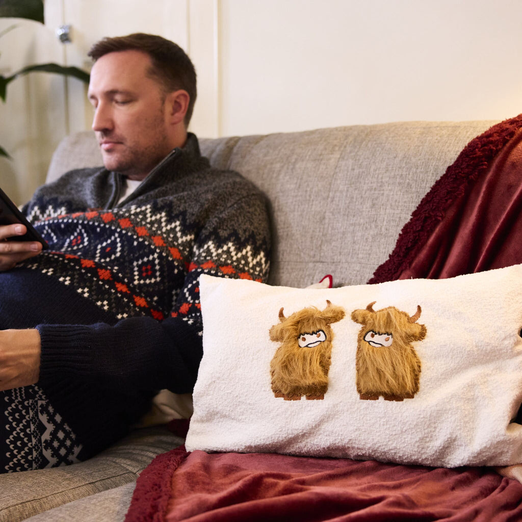 Man sits looking at his phone while the Highland Cow applique cushion is by his side.