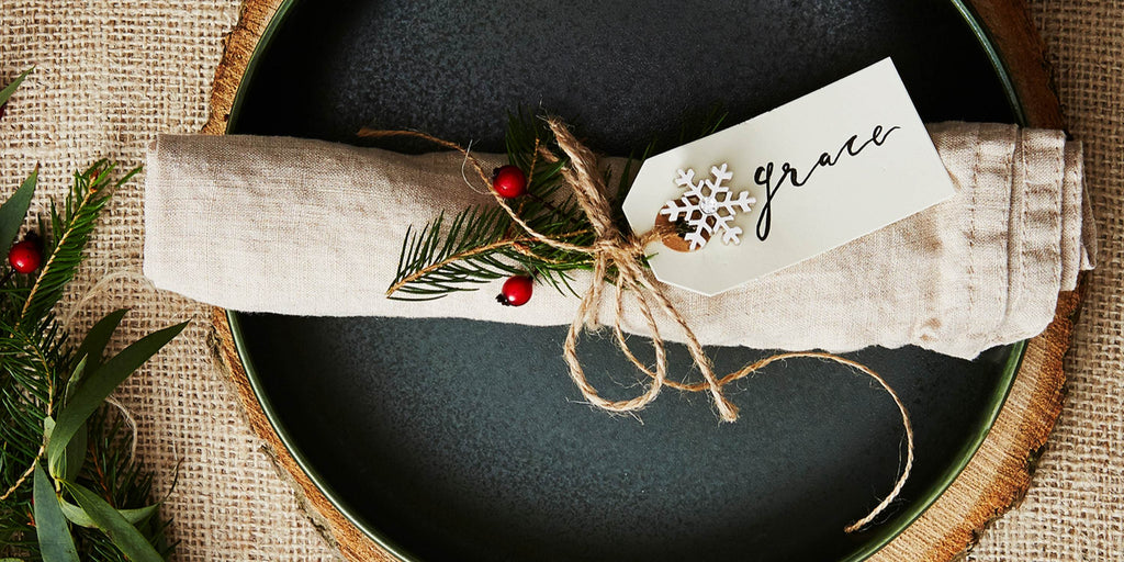 A snowflake-shaped pin badge is attached to a luggage label reading "Grace". It's tied, along with a sprig of pine, to a rolled up cotton napkin.