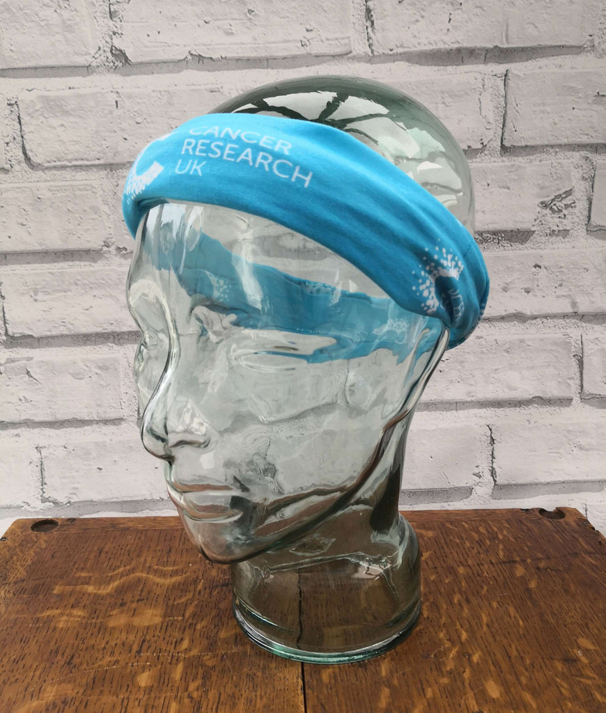 Blue multifunctional snood with Cancer Research pattern displayed on a mannequin head, styled as a forehead warmer. Ideal for post-chemo recovery, with soft, breathable fabric that won’t irritate sensitive skin.