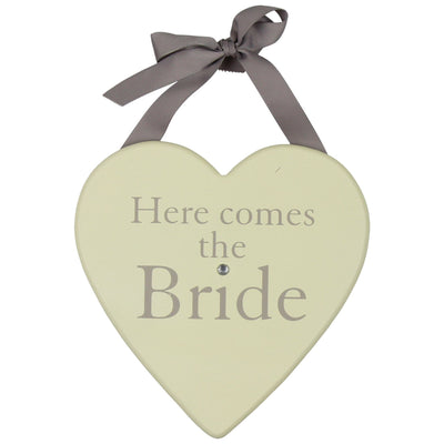 Heart-shaped decorative plaque in a soft cream colour with the words “Here comes the Bride” printed in pale grey lettering. A single small gem sits below the text. A grey ribbon is tied into a bow at the top for hanging. Presented as a special wedding keepsake.